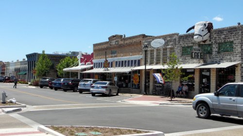 A charming street scene featuring shops, cafes, and parked cars under a clear blue sky. A cow sculpture adorns a building.