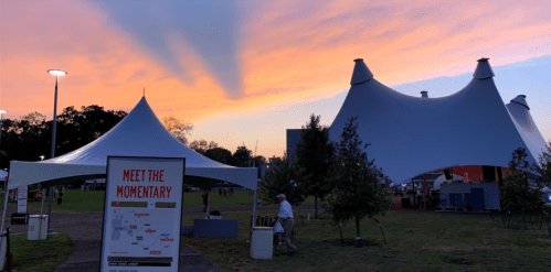 A sunset sky over a festival area with large tents and a sign reading "Meet the Momentary."