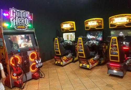 Arcade area featuring a Guitar Hero machine and racing game cabinets against a dark wall.