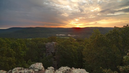 A scenic view of a sunset over rolling hills, with trees in the foreground and clouds in a colorful sky.