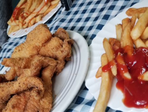 A plate of fried fish next to a plate of French fries drizzled with ketchup on a checkered tablecloth.
