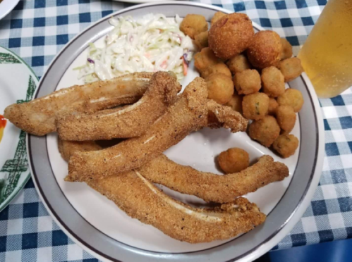 A plate of fried catfish fillets, hushpuppies, coleslaw, and fried okra on a checkered tablecloth.