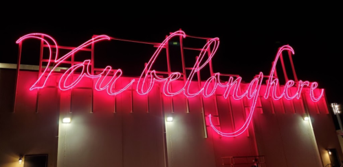A glowing pink neon sign reads "You belong here" against a dark night sky.