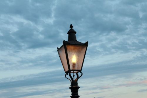 A vintage street lamp illuminated against a backdrop of cloudy blue sky.