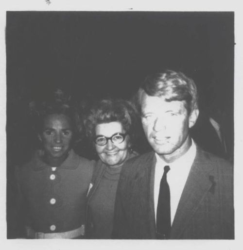 A black and white photo of three people smiling together, with one woman in a light coat and two others in formal attire.