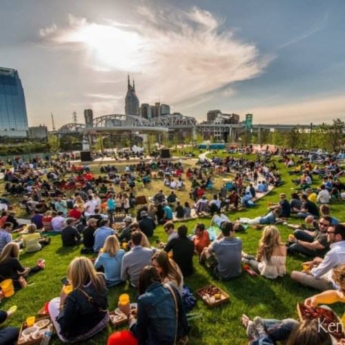 A large crowd enjoys a sunny outdoor event in a park, with a stage and city skyline in the background.