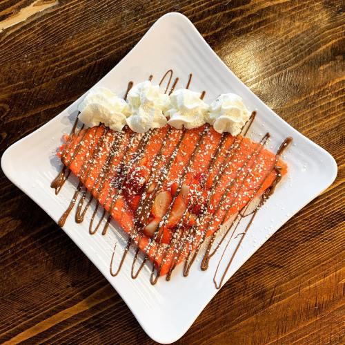 A heart-shaped dessert crepe topped with strawberries, whipped cream, and chocolate drizzle on a white plate.
