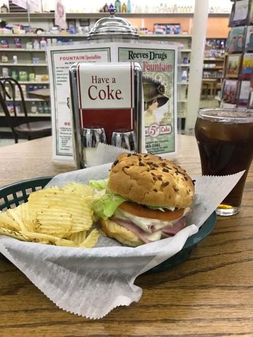 A sandwich with lettuce and ham, served with potato chips and a glass of cola, on a table in a retro drugstore.