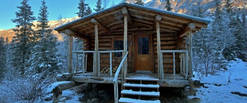 A cozy log cabin surrounded by snow-covered trees and mountains, with a wooden porch and steps leading to the door.