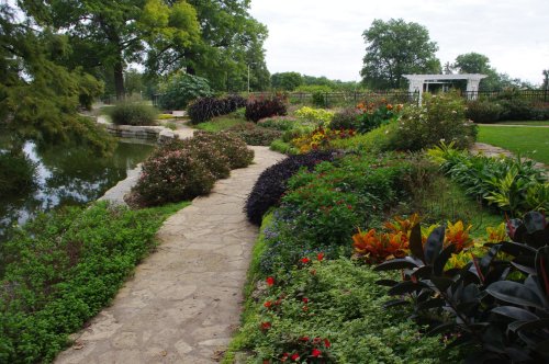 A scenic garden path lined with colorful flowers and greenery, leading to a tranquil pond and gazebo in the background.