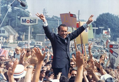A crowd cheers as a man in a suit raises his arms in victory, holding signs during a political rally.