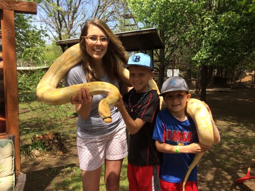 A woman and two boys hold a large yellow snake outdoors, surrounded by trees and a sunny backyard setting.