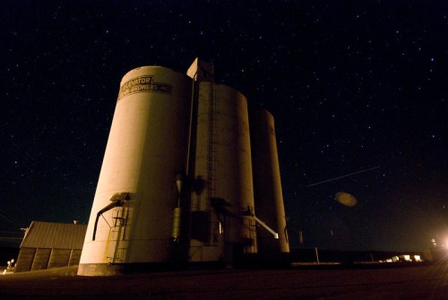 A large grain silo under a starry night sky, with faint lights in the background illuminating the scene.