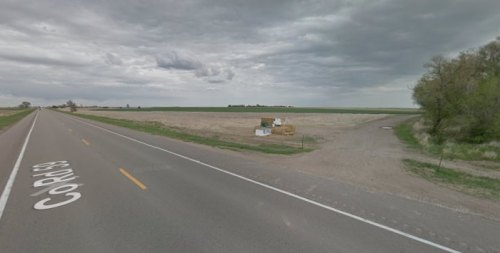 A rural road with a grassy area and a field under a cloudy sky, featuring a small structure and hay bales nearby.