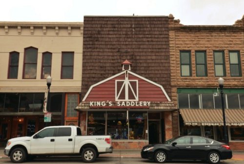 A storefront with a barn-style design, labeled "King's Saddlery," flanked by two parked cars on a city street.