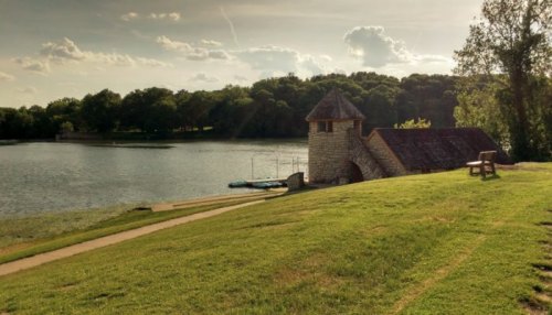 A serene lakeside scene featuring a stone building, grassy shore, and a bench under a cloudy sky.