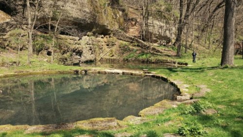 A serene pond surrounded by lush greenery and rocky cliffs, with a person walking along the water's edge.