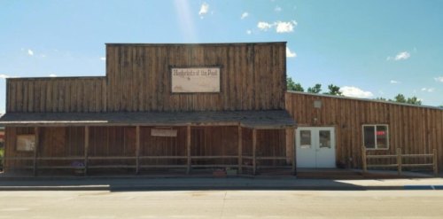 A rustic wooden building with a sign, featuring a porch and large windows, set against a clear blue sky.