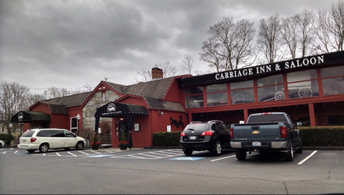 Exterior of Carriage Inn & Saloon, featuring a red building, large windows, and parked cars in front. Overcast sky.