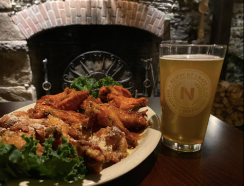 A plate of spicy chicken wings garnished with greens, alongside a glass of beer, with a brick fireplace in the background.