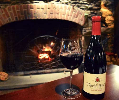 A glass of red wine beside a bottle of David Bruce Pinot Noir, with a cozy fireplace in the background.