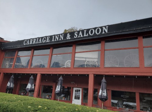Exterior of the Carriage Inn & Saloon, featuring large windows and umbrellas outside, with a red wooden facade.