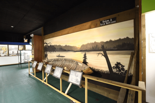 A mural depicting a sunset over a lake, with a birch bark canoe and framed images displayed in a gallery.