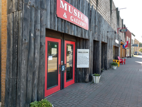 Entrance of a museum and gallery with red doors, wooden exterior, and potted plants along the sidewalk.