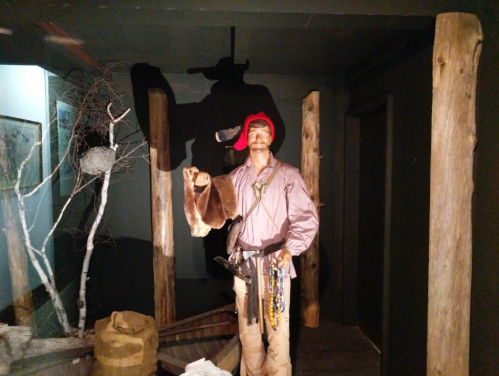 A life-sized figure of a frontiersman in period clothing, holding a fur and standing near wooden posts and barrels.