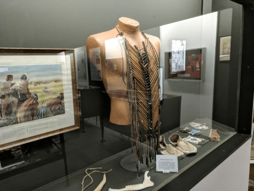 Display case featuring traditional Native American clothing and artifacts, including a beaded necklace and moccasins.