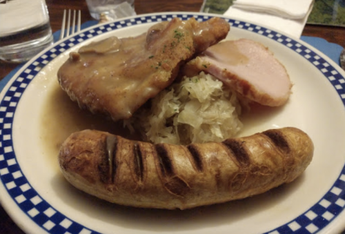 A plate of food featuring pork chops, sauerkraut, ham, and a grilled sausage, garnished with herbs.