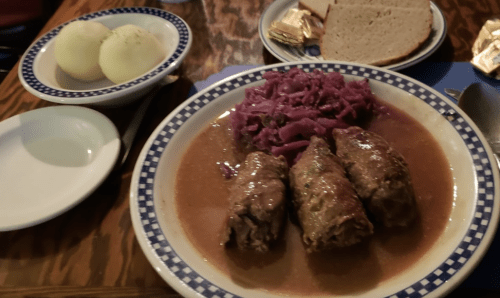 A plate of meatloaf with red cabbage and gravy, accompanied by boiled potatoes and slices of bread on a wooden table.
