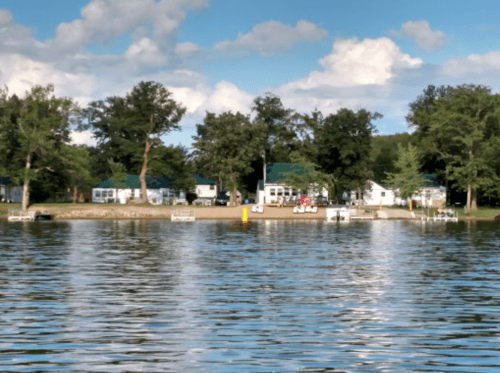 A serene lake view with cabins and trees along the shore under a blue sky with fluffy clouds.