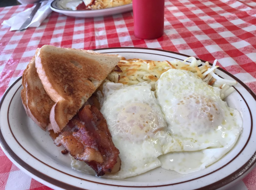 A plate of breakfast featuring two fried eggs, bacon, hash browns, and toast on a checkered tablecloth.