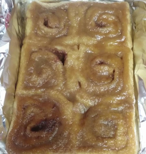 A tray of freshly baked cinnamon rolls, golden brown with a glossy glaze, arranged in a rectangular shape.