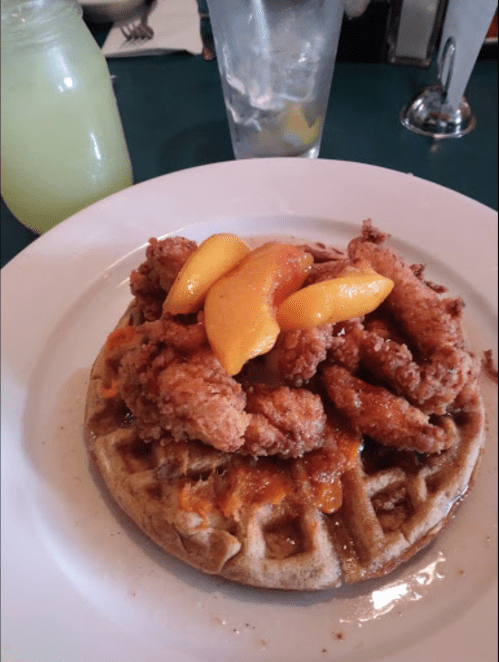 A plate featuring a waffle topped with fried chicken and peach slices, alongside a glass of lemonade.
