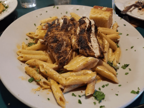 A plate of penne pasta topped with grilled chicken, broccoli, and a slice of cornbread, garnished with parsley.