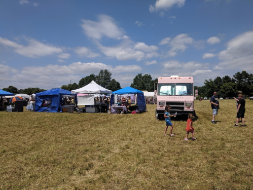 A vibrant outdoor market scene with tents, a food truck, and people, including children, enjoying the sunny day.
