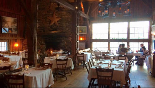 Cozy restaurant interior with wooden tables, chairs, and a stone fireplace, featuring warm lighting and rustic decor.