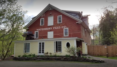 Red and yellow building with "Waterbury Feed Co." sign, surrounded by greenery and a gravel path.