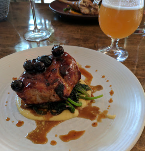 A plated dish featuring a glazed pork chop topped with blueberries, served on a bed of greens and a creamy sauce.
