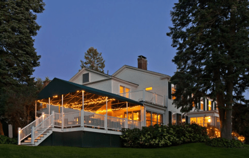 A charming white house with a green awning, illuminated by warm lights, surrounded by trees at dusk.