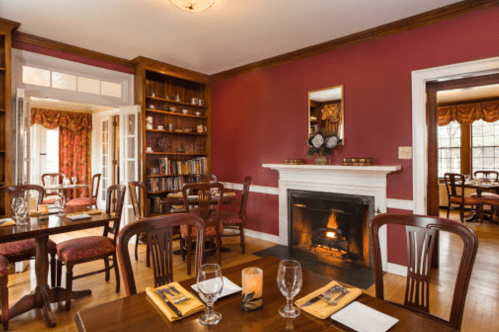 Cozy dining room with wooden tables, elegant chairs, a fireplace, and bookshelves, featuring warm red walls and soft lighting.