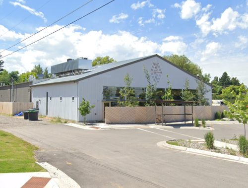 A modern, gray building with large windows, surrounded by greenery and a gravel area, under a blue sky with clouds.