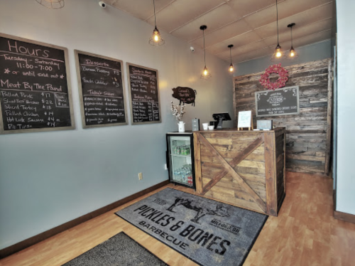 Interior of a barbecue restaurant with a wooden counter, menu boards, and a welcome mat reading "Pickles & Bones Barbecue."