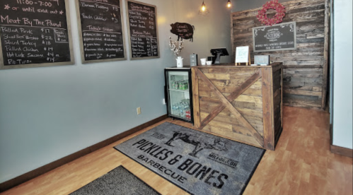 Interior of a barbecue restaurant with a wooden counter, menu boards, and a welcome mat that reads "Pickles & Bones Barbecue."