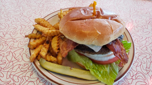 A juicy burger with bacon, lettuce, tomato, and a pickle, served with crinkle-cut fries on a patterned table.