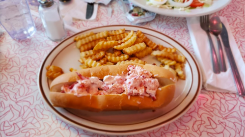A plate with a lobster roll filled with creamy lobster and a side of crinkle-cut fries.