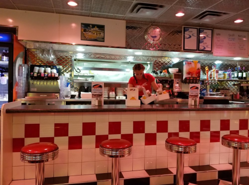A diner counter with red and white tiles, featuring a server preparing food behind the counter. Retro decor is visible.