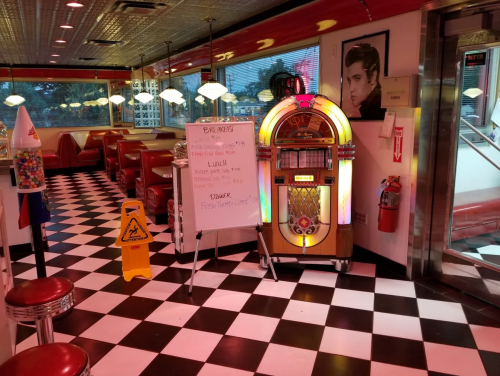 A retro diner with red booths, a jukebox, and a checkered floor, featuring a menu board and a caution sign.